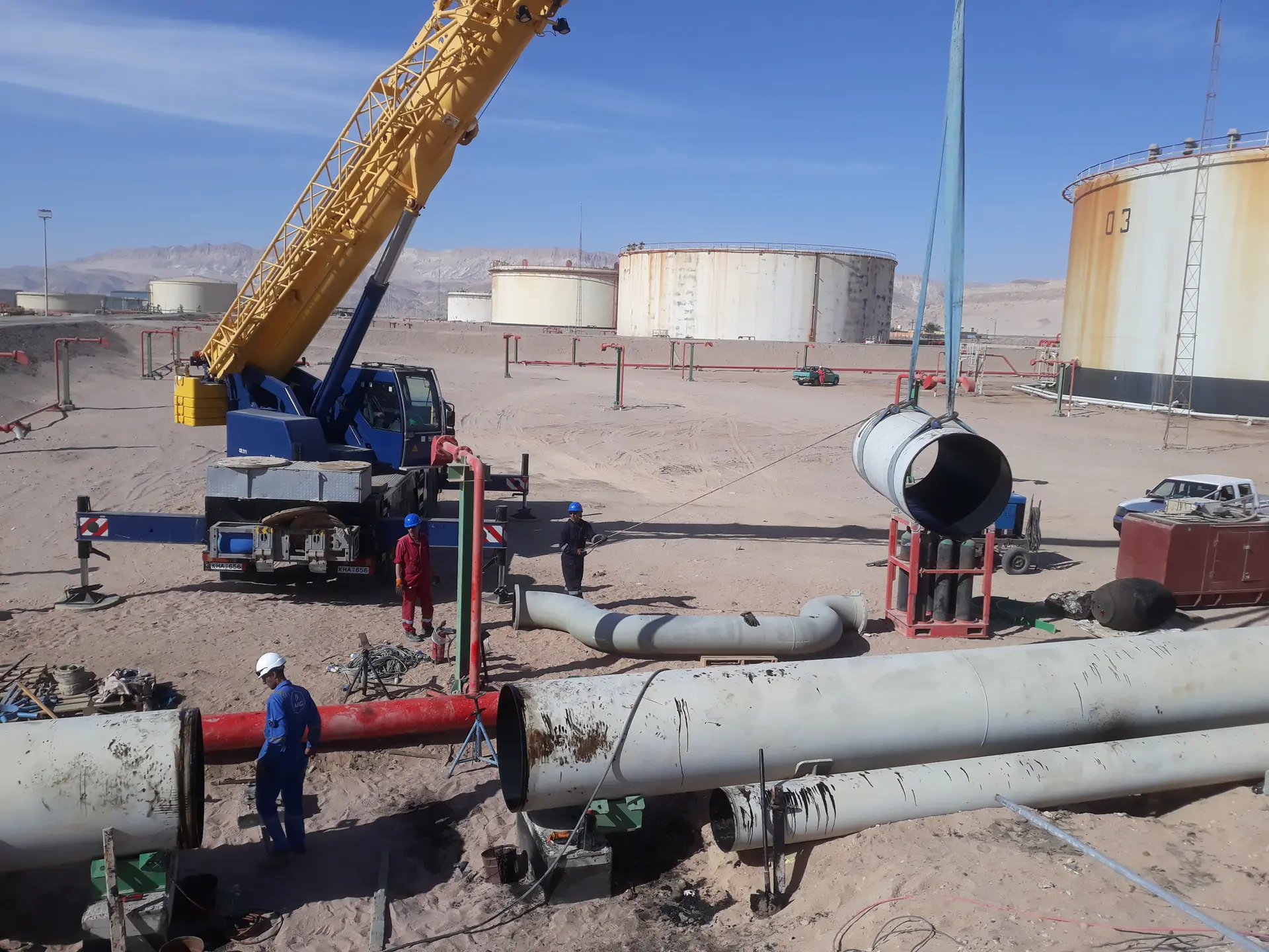 Mobile crane lifting a pipe spool beside storage tanks